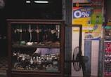A display cabinet filled with a variety of wristwatches, including those with leather and metal bands. Behind the glass display, there is a person partially visible, wearing a white shirt. On the adjacent wall, colorful advertisements and signs are visible, and a standing fan is placed next to the cabinet.