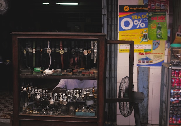 A display cabinet filled with a variety of wristwatches, including those with leather and metal bands. Behind the glass display, there is a person partially visible, wearing a white shirt. On the adjacent wall, colorful advertisements and signs are visible, and a standing fan is placed next to the cabinet.