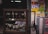 A display cabinet filled with a variety of wristwatches, including those with leather and metal bands. Behind the glass display, there is a person partially visible, wearing a white shirt. On the adjacent wall, colorful advertisements and signs are visible, and a standing fan is placed next to the cabinet.