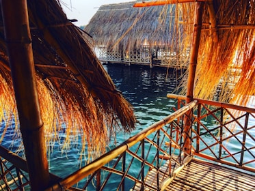 A view of a wooden and bamboo hut constructed over water, with thatched roofs and walls. The structure is built with interwoven bamboo patterns, and the water beneath is a vibrant blue-green. Sunlight streams through, casting warm, golden hues on the bamboo.