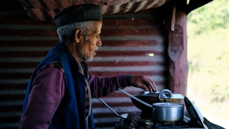 An elderly man is cooking in a rustic kitchen. He wears a traditional cap and layered clothing, standing next to a stove with pots. The background features a textured wall and wooden beams, creating a warm and homely atmosphere.