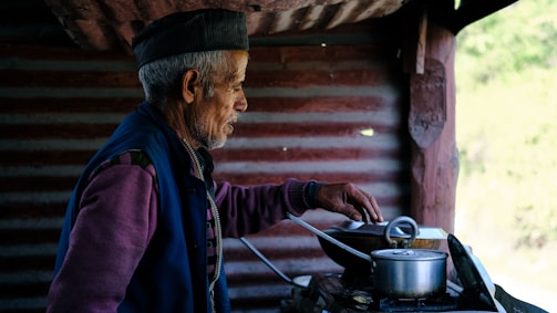 An elderly man is cooking in a rustic kitchen. He wears a traditional cap and layered clothing, standing next to a stove with pots. The background features a textured wall and wooden beams, creating a warm and homely atmosphere.