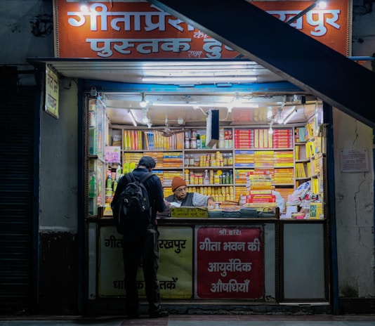 A small bookshop with bright fluorescent lighting, crammed with neatly arranged stacks of books and products. The exterior has a sign in Hindi, and there are two individuals at the counter, one browsing and the other, wearing a beanie, assisting. The shop is painted in warm tones, creating a cozy atmosphere.