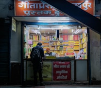 A small bookshop with bright fluorescent lighting, crammed with neatly arranged stacks of books and products. The exterior has a sign in Hindi, and there are two individuals at the counter, one browsing and the other, wearing a beanie, assisting. The shop is painted in warm tones, creating a cozy atmosphere.