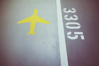 A section of pavement features a bright yellow airplane symbol to the left and the numbers 3305 in white on the right. A white line separates these elements on a textured surface.