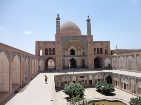 An Islamic architectural structure with an expansive courtyard and a large central dome, adorned with intricate tile work. The building features multiple arches and minarets, surrounded by high walls with geometric patterns. People are scattered around the courtyard, and a small garden is present in the center.