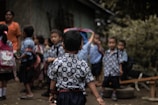 A group of young children wearing identical patterned shirts are gathered outdoors. They appear to be preparing for or returning from school. Some children have backpacks, and they stand on what looks like a dirt path surrounded by greenery. An adult stands nearby, wearing an orange shirt.