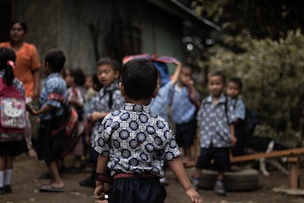 A group of young children wearing identical patterned shirts are gathered outdoors. They appear to be preparing for or returning from school. Some children have backpacks, and they stand on what looks like a dirt path surrounded by greenery. An adult stands nearby, wearing an orange shirt.