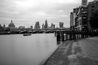 A black and white photograph of a riverside cityscape featuring a prominent cathedral dome, modern skyscrapers, and a bridge spanning a calm river. The foreground includes a pebble-strewn shoreline and wooden piers, with buildings and the riverbank stretching into the distance.