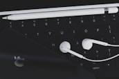 An arrangement of technological accessories featuring a keyboard, white earphones, a white stylus, and a smartphone with a visible logo on a dark background.
