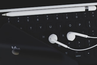 An arrangement of technological accessories featuring a keyboard, white earphones, a white stylus, and a smartphone with a visible logo on a dark background.
