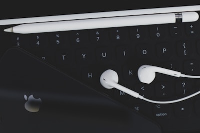 An arrangement of technological accessories featuring a keyboard, white earphones, a white stylus, and a smartphone with a visible logo on a dark background.