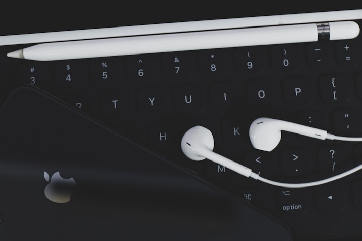 An arrangement of technological accessories featuring a keyboard, white earphones, a white stylus, and a smartphone with a visible logo on a dark background.