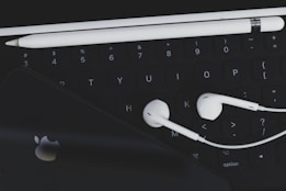 An arrangement of technological accessories featuring a keyboard, white earphones, a white stylus, and a smartphone with a visible logo on a dark background.