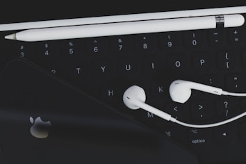 An arrangement of technological accessories featuring a keyboard, white earphones, a white stylus, and a smartphone with a visible logo on a dark background.