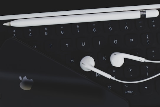 An arrangement of technological accessories featuring a keyboard, white earphones, a white stylus, and a smartphone with a visible logo on a dark background.