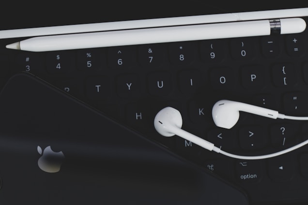 An arrangement of technological accessories featuring a keyboard, white earphones, a white stylus, and a smartphone with a visible logo on a dark background.