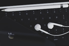 An arrangement of technological accessories featuring a keyboard, white earphones, a white stylus, and a smartphone with a visible logo on a dark background.
