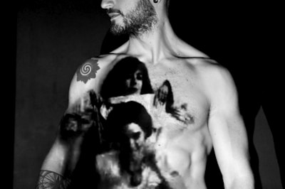 A shirtless person with tattoos on their arm and chest is featured. A black and white artistic projection of multiple human faces is cast across their torso, creating a striking contrast against their skin. The overall composition is dramatic, with strong shadows and highlights enhancing the mood.