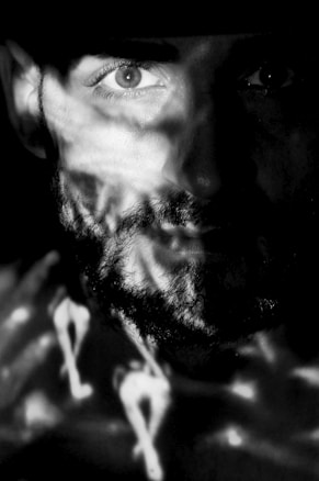 An artistic black and white portrait of a bearded person. Partial illumination and shadow patterns create a sense of mystery and focus on their eye. The use of light and shadow adds an abstract quality to the image.