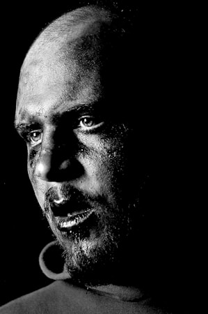 A black and white portrait captures a man's face in dramatic lighting. The high contrast highlights the texture of the skin and the intense expression, particularly around the eyes.