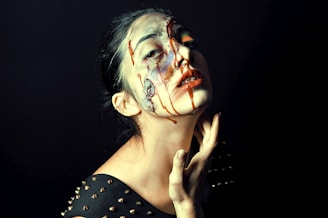 A person with dramatic and expressive makeup has colorful paint and what appears to be dark red liquid running down their face. Their eyes are partially closed, and the expression hints at passion or pain. The background is dark, adding contrast and drama to the image.