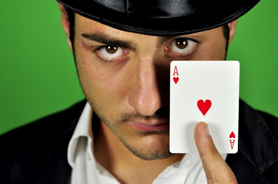 A person wearing a black hat and suit holds an Ace of Hearts playing card close to their face, against a green background. The expression is intense and focused, with the card prominently displayed.