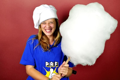 A person is smiling broadly while holding a large stick of cotton candy. They are wearing a blue shirt with a logo and a white chef's hat. The background is a solid red color.