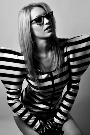 A person with long hair wearing glasses and a striped top with exaggerated shoulders poses in a fashion-forward style. The image is in black and white, enhancing the boldness of the outfit's geometric pattern.