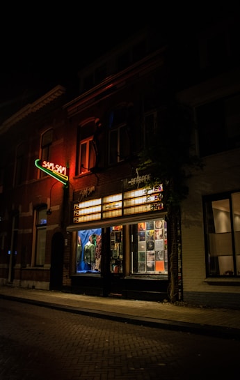 A dimly lit street scene at night featuring a brick building with illuminated signage indicating 'Sam Sam.' The building has large windows displaying various items, possibly including clothing and posters, with a warm, amber glow coming from interior lighting.