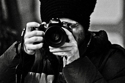 A person wearing a dark jacket and a beanie is holding a Nikon DSLR camera close to their face, ready to take a photograph. The image is in black and white, adding a classic, timeless feel.