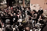 A group of people gathered outdoors, engaging in conversation or listening intently. They are dressed warmly, indicating a cold day. The background shows trees and a wall with graffiti, suggesting an urban setting.