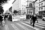A large group of people is participating in a street protest, holding a banner with Italian writing. They are walking down a cobblestone street surrounded by multi-story buildings with a prominent statue. The atmosphere seems lively and determined.