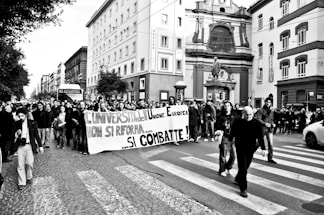 A large group of people is participating in a street protest, holding a banner with Italian writing. They are walking down a cobblestone street surrounded by multi-story buildings with a prominent statue. The atmosphere seems lively and determined.