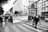 A large group of people is participating in a street protest, holding a banner with Italian writing. They are walking down a cobblestone street surrounded by multi-story buildings with a prominent statue. The atmosphere seems lively and determined.