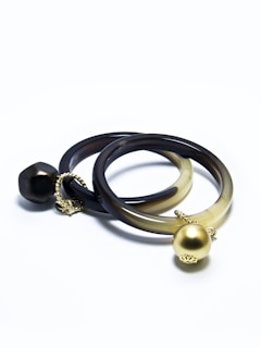 Two intertwined bracelets with a smooth, dark finish. Each bracelet features a distinct decorative element: one with a large gold spherical charm, the other adorned with a unique darker bead. Both are connected by small gold chains, creating a sophisticated and elegant look.