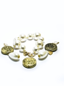 A bracelet composed of large, glossy white pearls and ornate gold coins, interspersed with teardrop-shaped white beads. The gold coins feature intricate engravings, adding a touch of elegance. The pearls and gold elements are linked together in a cohesive, stylish design.