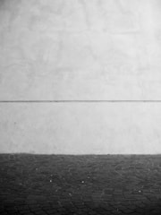 A minimalist black and white composition featuring a textured stone pavement extending to a plain wall, divided by a subtle horizontal line.