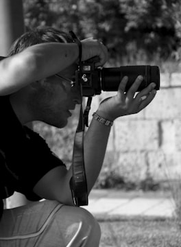 A person is crouched down, focusing through the viewfinder of a camera with a large lens. The setting appears to be outdoors with a blurred background. The person is wearing a bracelet and glasses, and their expression is concentrated.