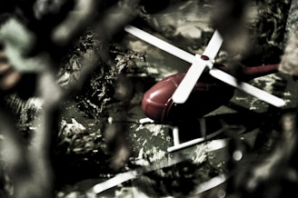 A close-up of a small red toy helicopter with white blades, surrounded by blurred foliage in a seemingly outdoor setting. The helicopter is on a surface that appears to mimic a natural environment, possibly a toy model setup.