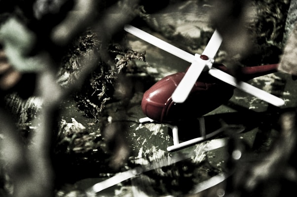 A close-up of a small red toy helicopter with white blades, surrounded by blurred foliage in a seemingly outdoor setting. The helicopter is on a surface that appears to mimic a natural environment, possibly a toy model setup.
