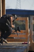 A person wearing protective gear is crouched behind wooden barricades, holding a paintball gun. The scene takes place outdoors with a structure and clear sky in the background.