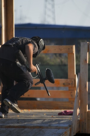 A person wearing protective gear is crouched behind wooden barricades, holding a paintball gun. The scene takes place outdoors with a structure and clear sky in the background.