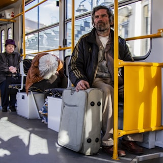 Several people are seated inside a public transportation vehicle. A middle-aged man in a dark jacket holding a suitcase is sitting on the right side. Another person with white hair is sitting hunched over to the left, and an older woman wearing a purple hat is seated further back. The atmosphere appears quiet and pensive.