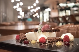 A close-up of a gourmet dessert plate featuring a mix of textures and colors. There are pieces of brownie, a scoop of berry sorbet, fresh raspberries, dollops of cream, and decorative sauces, all arranged artistically. The background is a softly lit, out-of-focus restaurant interior with warm tones and hanging lights creating a bokeh effect.