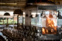 The image features a cozy indoor setting with a fireplace prominently displaying bright, warm flames. The background is softly blurred, suggesting a rustic dining or living area with tables and chairs. Light filters through large windows, creating a pleasant, inviting atmosphere.