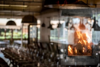 The image features a cozy indoor setting with a fireplace prominently displaying bright, warm flames. The background is softly blurred, suggesting a rustic dining or living area with tables and chairs. Light filters through large windows, creating a pleasant, inviting atmosphere.