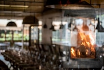 The image features a cozy indoor setting with a fireplace prominently displaying bright, warm flames. The background is softly blurred, suggesting a rustic dining or living area with tables and chairs. Light filters through large windows, creating a pleasant, inviting atmosphere.