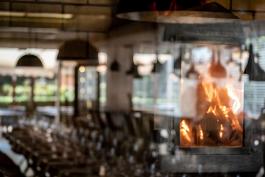 The image features a cozy indoor setting with a fireplace prominently displaying bright, warm flames. The background is softly blurred, suggesting a rustic dining or living area with tables and chairs. Light filters through large windows, creating a pleasant, inviting atmosphere.