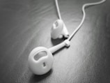 A close-up view of a pair of white earbud headphones resting on a textured, dark surface. The earbuds have a sleek and minimalist design with slight reflections on the surface, suggesting a smooth finish.